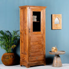 Side angle image of a antique teak wood display cabinet with glass door, shelves, and lower drawer – unique furniture piece crafted from teak wood with traditional Indian design and honey wax finish.