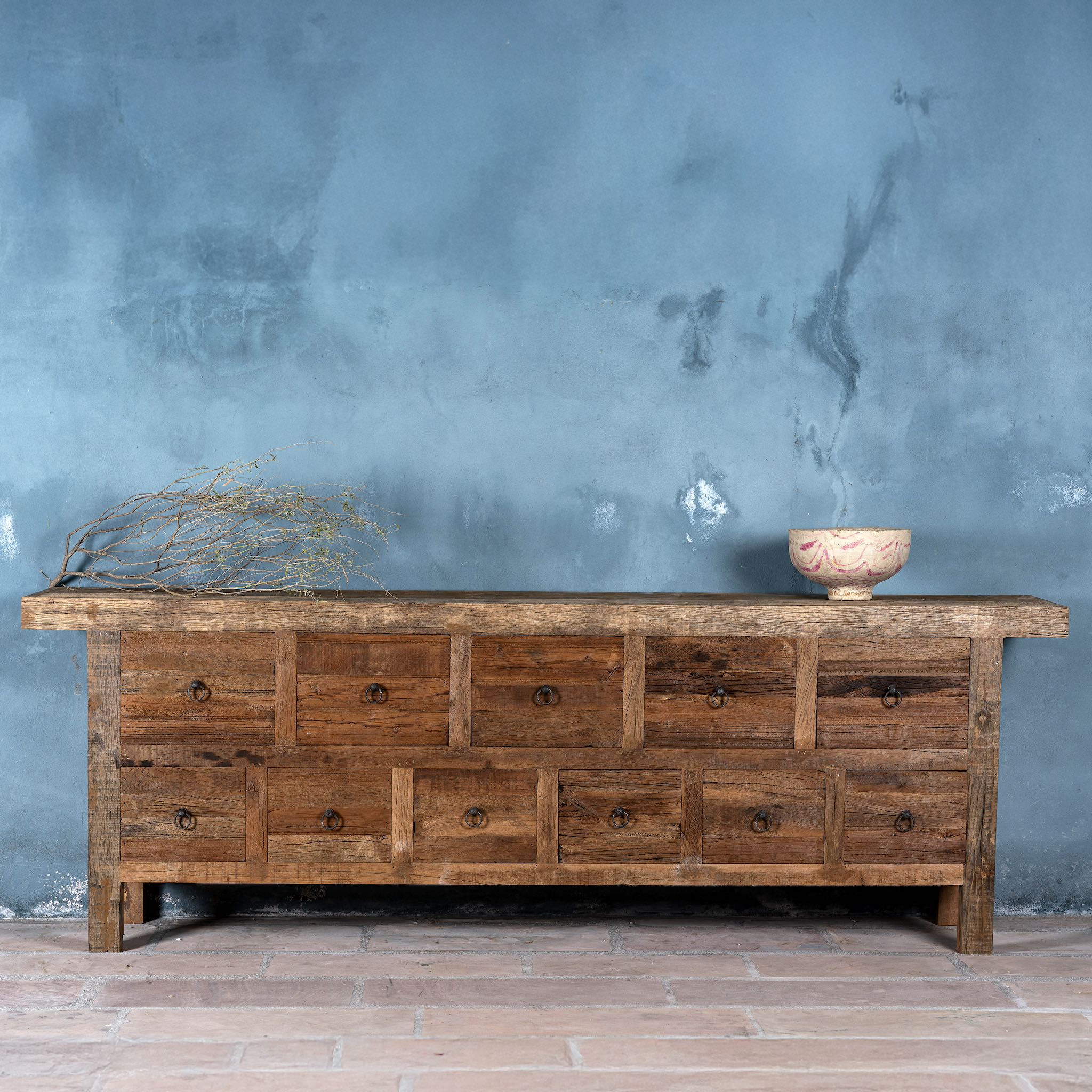 Front view image of a large wabi sabi style console table crafted from reclaimed wood, featuring a natural weathered finish and multiple drawers with simple metal ring pulls, embracing rustic imperfection and minimalist charm.
