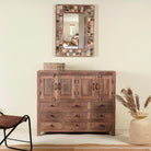 An elegant teak wood sideboard with multiple drawers and cupboards, featuring a brown finish. It is placed against a wall with a decorative mirror above it, and there is a vase with dried plants to the side.