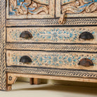 Close up image of the leg part of a hand-painted cabinet made from mango wood, featuring rattan inlay doors framed by a carved circular motif, floral blue and green accents, and five drawers for additional storage.