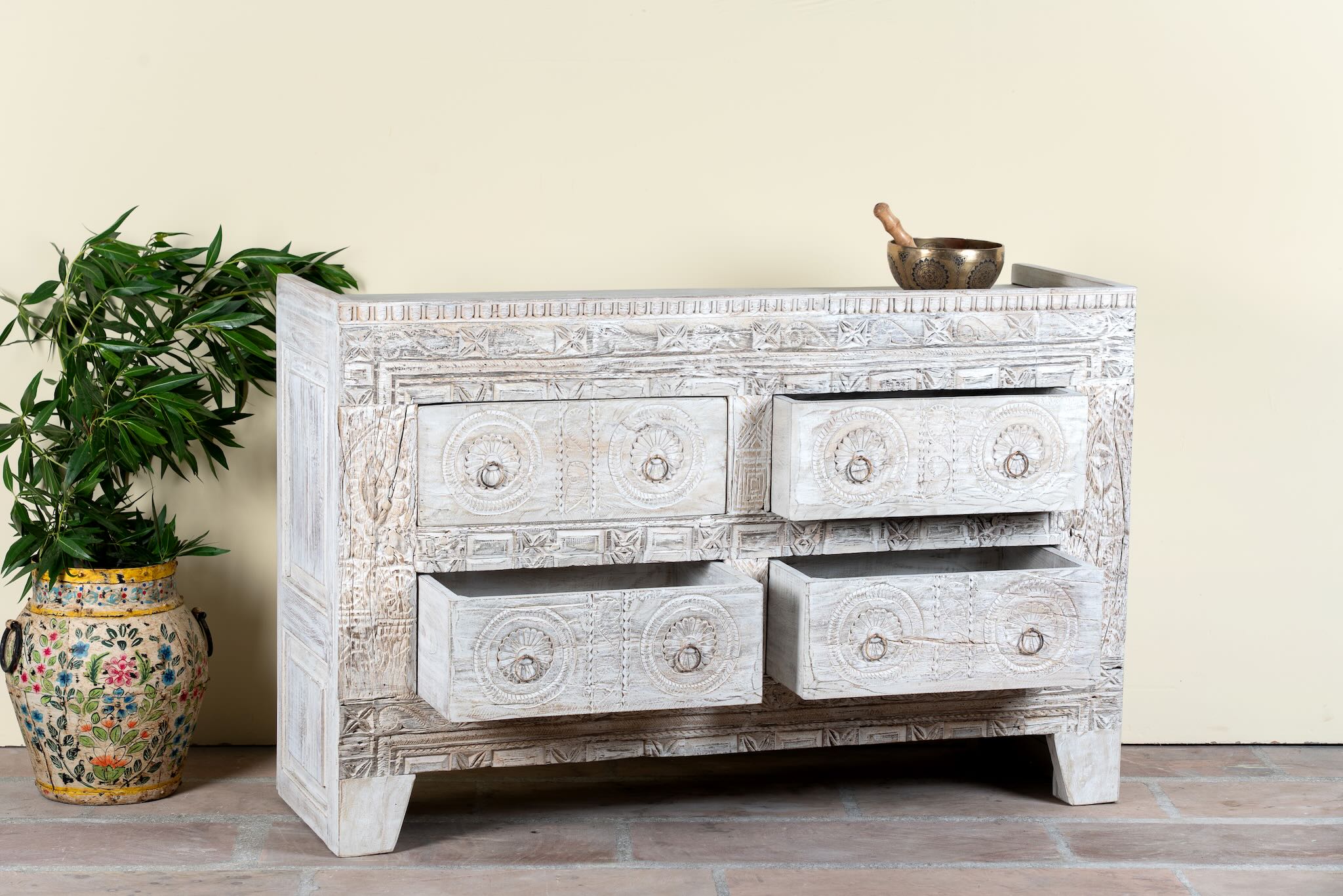 White distressed dresser with decorative carvings on a wooden floor with a plant and vase in the background.