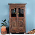 A tall, refurbished antique teak wood showcase with a glass front and a bird perched on a decorative element, placed against a light blue wall with a brown plant beside it.