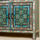Close up image of the leg part of a hand-painted reclaimed wood sideboard with floral and geometric patterns in vibrant hues. The sideboard has multiple drawers and cabinet doors, and is placed in a room with a vase on top and a chair to the side.