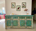 Lifestyle image of a hand-painted reclaimed wood sideboard with floral and geometric patterns in vibrant hues. The sideboard has multiple drawers and cabinet doors, and is placed in a room with a vase on top and a chair to the side.