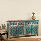 Side angle image of a hand-painted reclaimed wood sideboard with floral and geometric patterns in vibrant hues. The sideboard has multiple drawers and cabinet doors, and is placed in a room with a vase on top and a chair to the side.