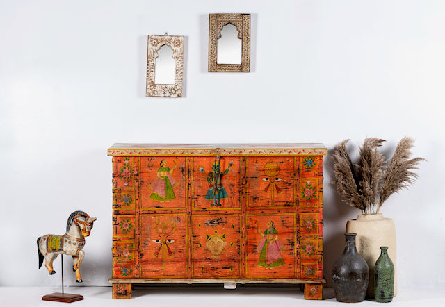 Hand painted teak wood storage chest with vibrant orange color and traditional Indian patterns, displayed on a white background with decorative items around it.