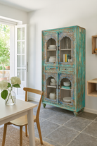 Lifestyle image of a hand-carved mango wood showcase with glass doors and drawers – carved wooden furniture in distressed turquoise and blue, combining artisan detail with vintage charm.