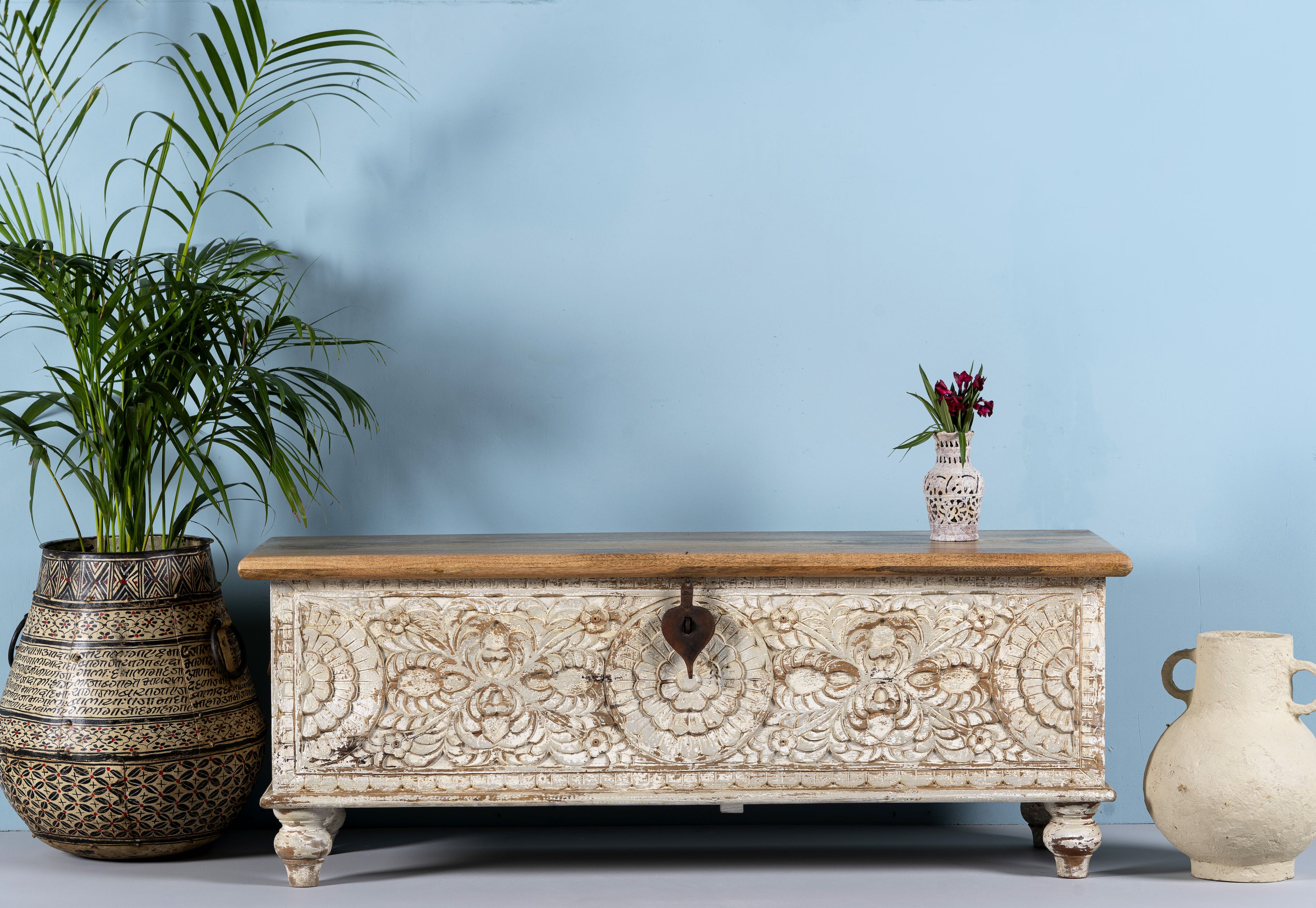 Hand-carved mango wood storage box with floral patterns and a natural wood top, accompanied by a vase with flowers and a pot with a plant.