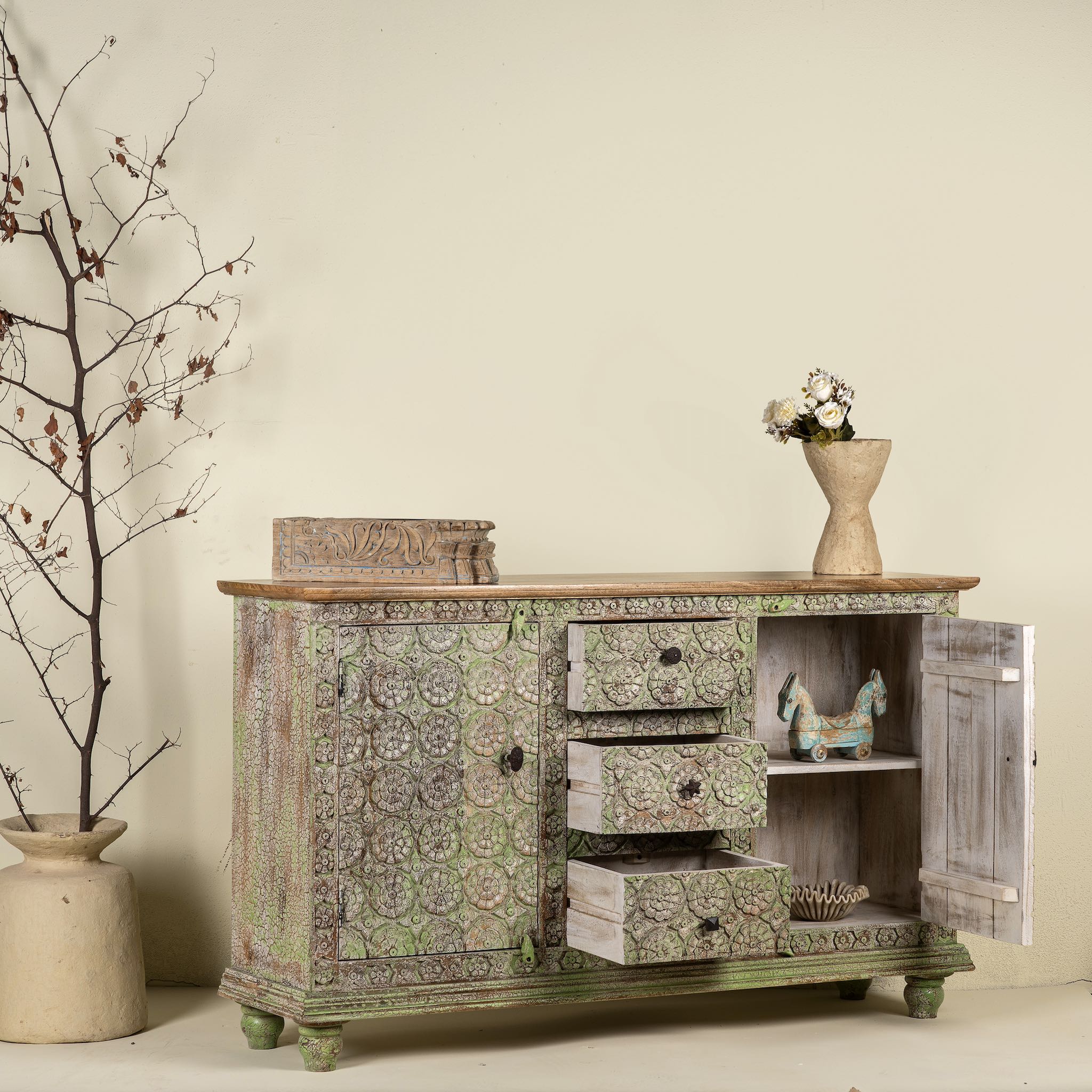 Side angle image with open door of a carved wooden furniture sideboard with floral hand-carving and distressed finish, vintage style cabinet made from mango wood, 150 × 40 × 90 cm.