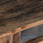 Close up picture of a rustic wooden bench made from reclaimed wood, featuring a long, narrow seat with a weathered finish and sturdy angled legs connected by a simple support beam.

