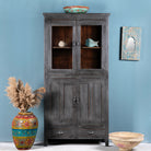 A refurbished teak wood showcase with glass doors and multiple compartments, featuring a shabby chic grey finish. It is positioned against a blue wall with decorative vases and artwork behind it.