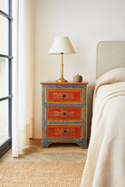 Lifestyle scene with a hand-painted reclaimed wood nightstand with vibrant floral motifs, featuring three drawers and a recycled wood top, placed next to a vase with dried plants.
