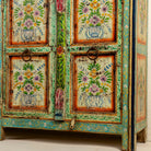 Close up image of the leg part of a hand-painted sideboard made from reclaimed wood, showcasing vibrant floral motifs in green, pink, yellow, and blue on a distressed turquoise base, with six panelled doors and rustic iron ring handles.