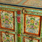 Close up image of a hand-painted sideboard made from reclaimed wood, showcasing vibrant floral motifs in green, pink, yellow, and blue on a distressed turquoise base, with six panelled doors and rustic iron ring handles.