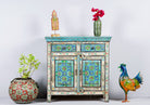 A hand-painted vintage cupboard with floral and geometric patterns in vibrant shades of blue, green, and red, on a distressed off-white base. The cupboard has two drawers and two doors.
