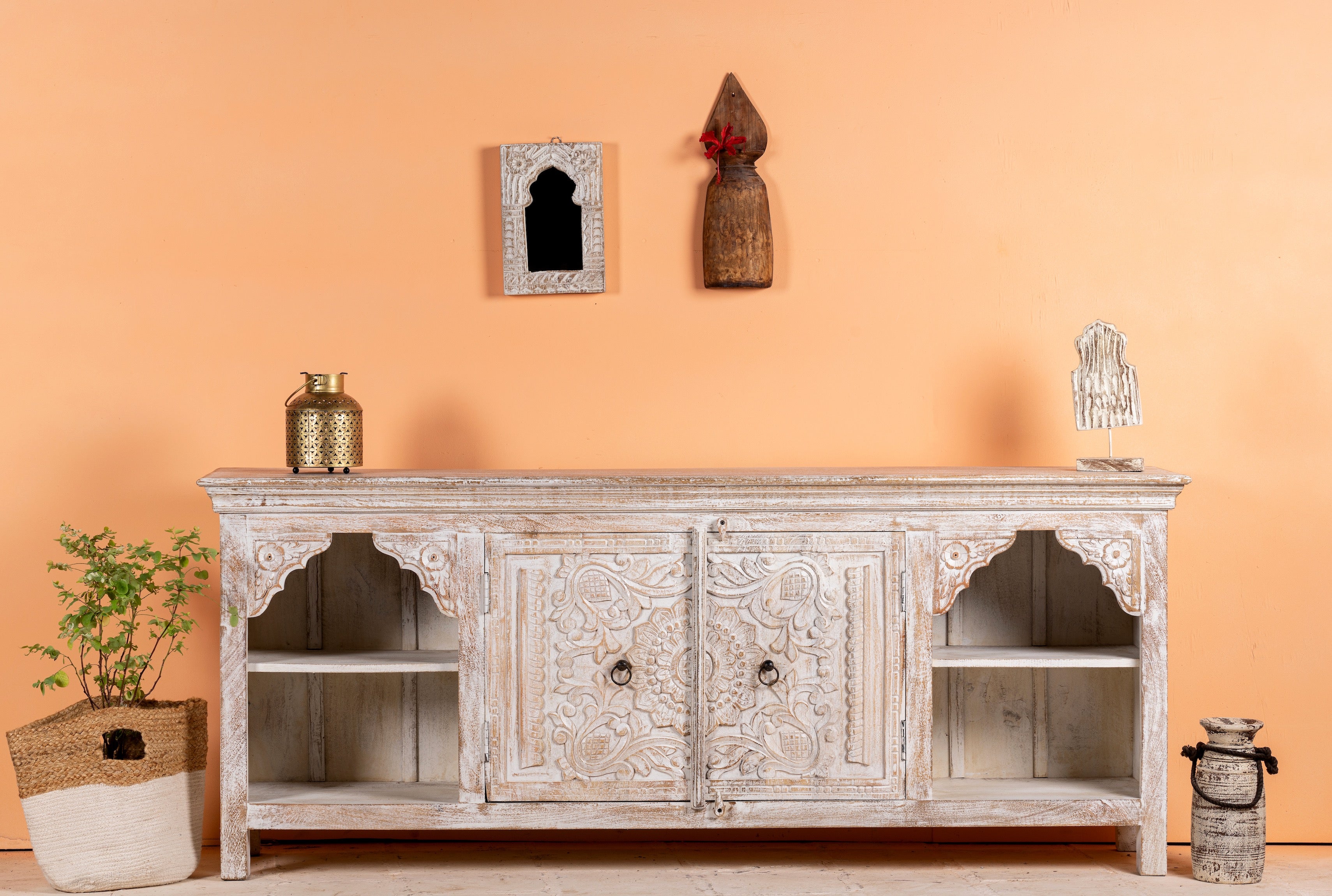 A white washed handcrafted Indian sideboard 'BHESAN' made from mango wood, featuring floral engravings, placed against an orange wall with decorative items on top and sides.
