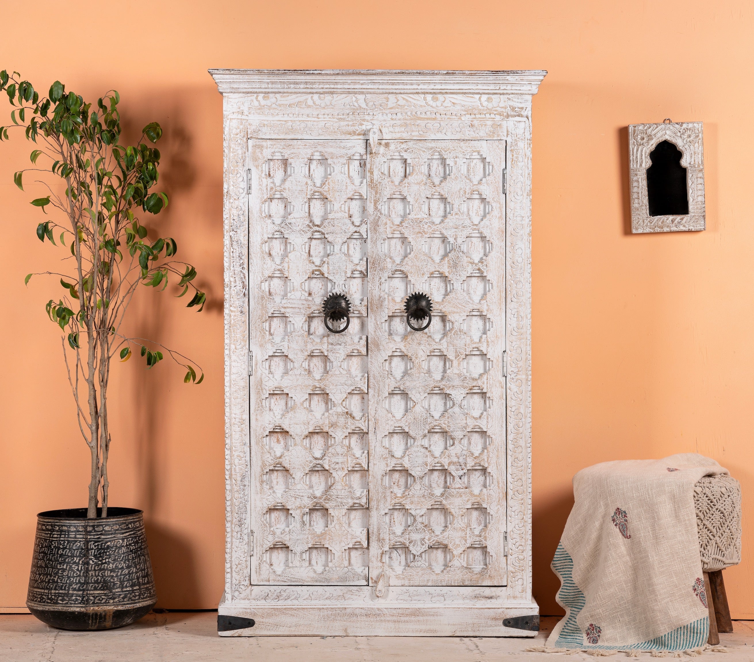 A white-washed handcrafted cabinet made from reclaimed wood, featuring intricate hand carvings and iron fittings, set against an orange wall with a potted plant to the side.
