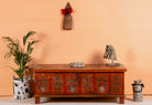 A hand-painted wooden box table with floral patterns, placed against an orange backdrop with decorative items on and around it, including a potted plant and a sculpture.