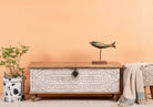 A hand-carved mango wood storage box with intricate patterns and antique-style iron fittings, finished in a distressed white, placed in a room with a plant and a decorative fish figure on top.