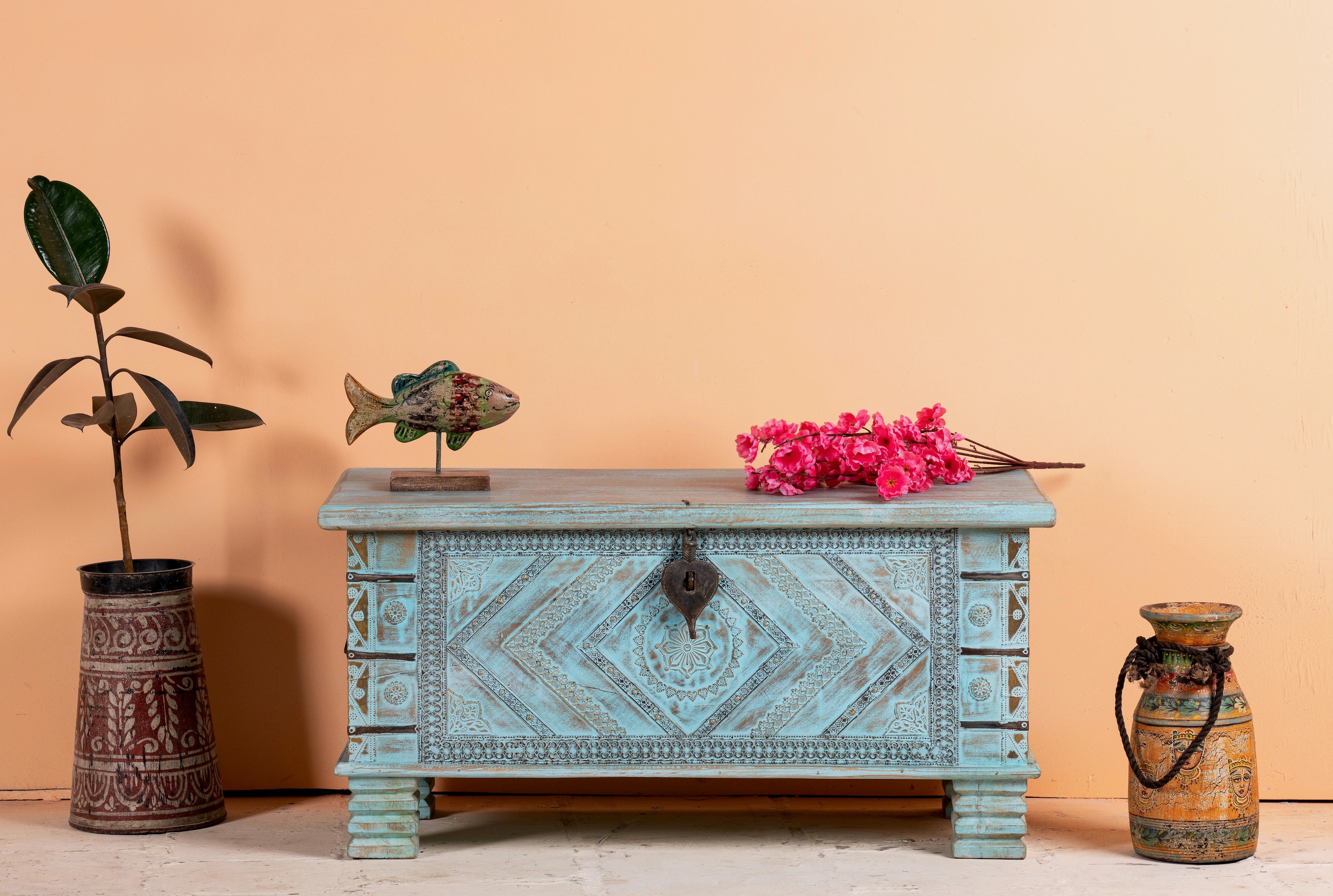 A hand-carved blue mango wood storage box with iron fittings, placed against a neutral background with a plant and decorative items on and around it.