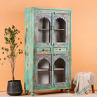 Side angle image of a hand-carved mango wood showcase with glass doors and drawers – carved wooden furniture in distressed turquoise and blue, combining artisan detail with vintage charm.