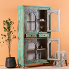 Side angle image with open door of a hand-carved mango wood showcase with glass doors and drawers – carved wooden furniture in distressed turquoise and blue, combining artisan detail with vintage charm.