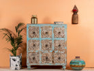 A vintage design mango wood chest of nine drawers with hand-carved floral patterns on the drawer fronts, featuring a blue body with whitewashed detailing and iron handles. The chest is positioned against a pink wall with a potted plant and decorative items on either side.