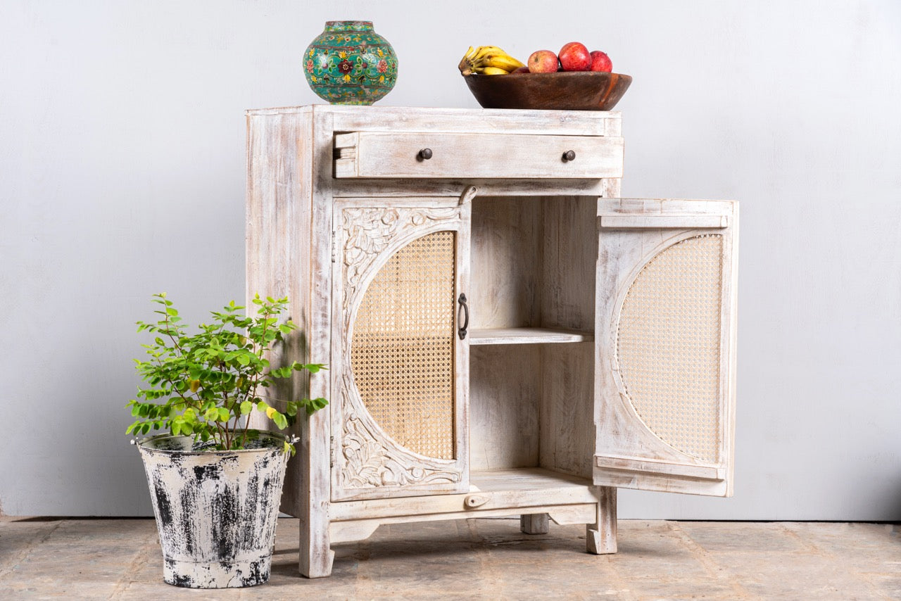Side angle image with open door of a white‑washed mango wood cupboard with rattan door panels and hand‑carved Indian ornamentation, 80 × 40 × 104 cm.